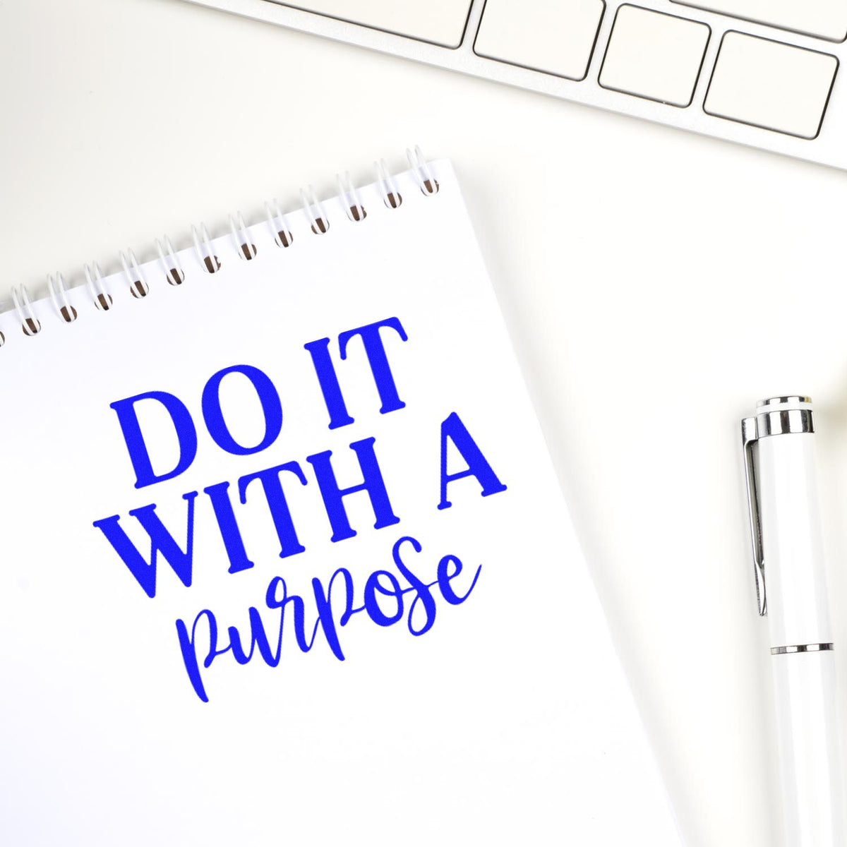 Blue custom stamp design reading Do It With a Purpose on a white spiral notebook, with a white pen and keyboard.