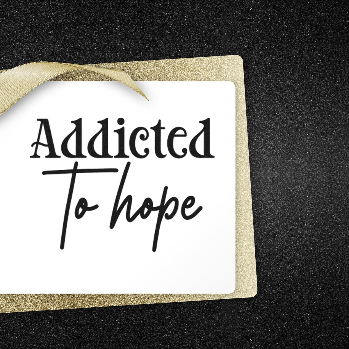 Custom stamp and seal reading Addicted to hope on a white card with gold glitter border and ribbon, on a black background.