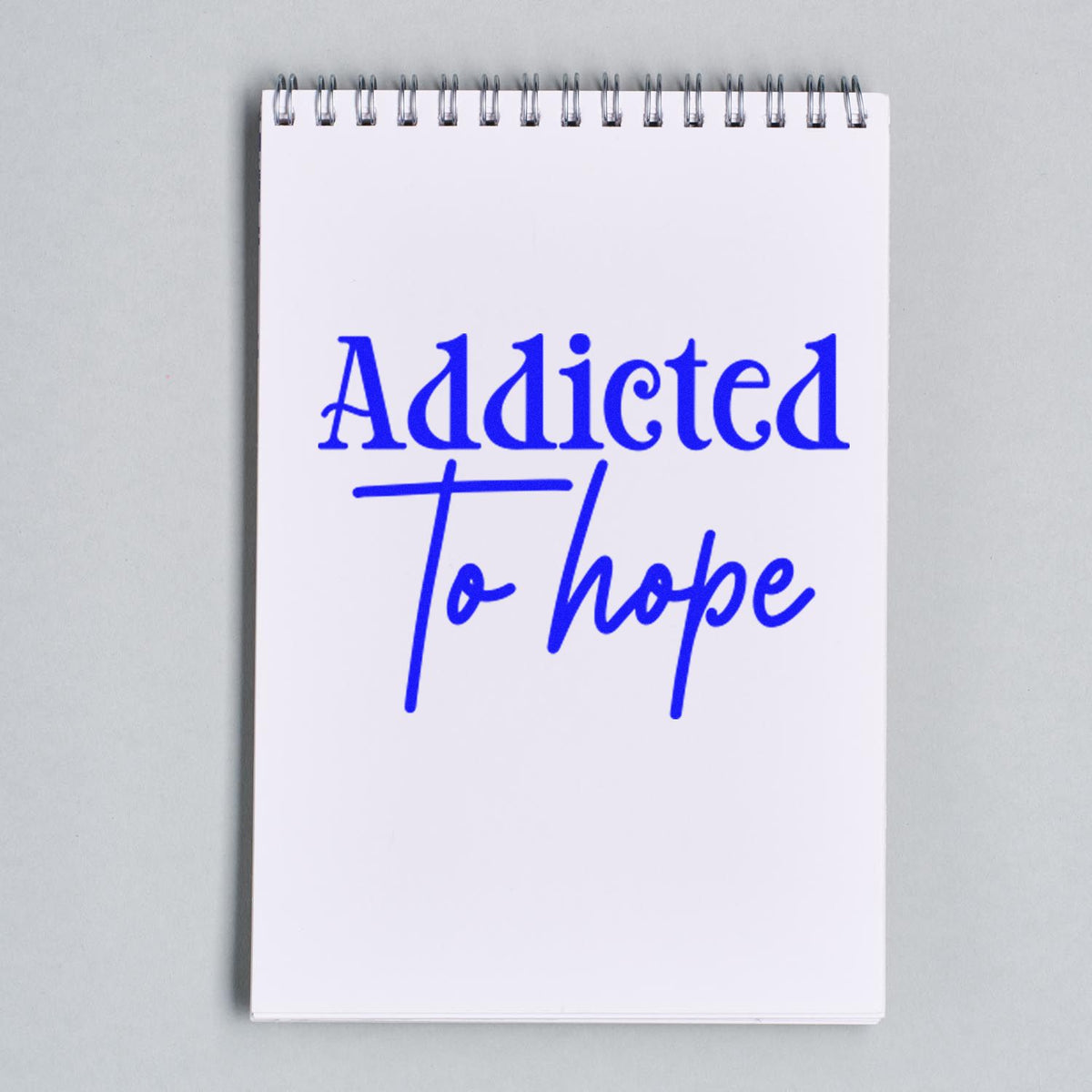 Blue stamp and seal imprint reading Addicted To Hope on a white spiral notebook.