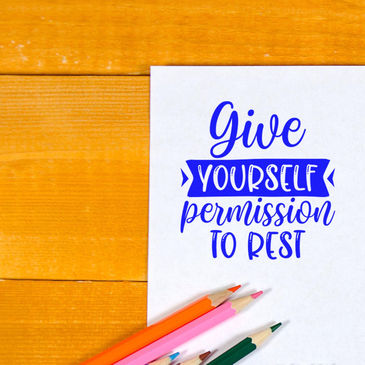 Blue stamp-and-seal design on white card: Give yourself permission to rest, with colored pencils on warm wood.