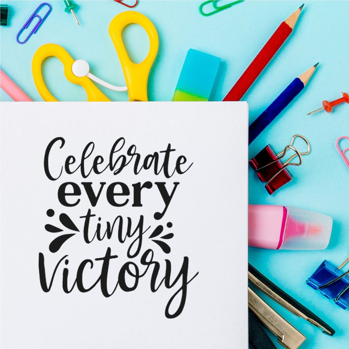 White stamp and seal card reading Celebrate every tiny victory on aqua background with colorful clips, pencils, and eraser