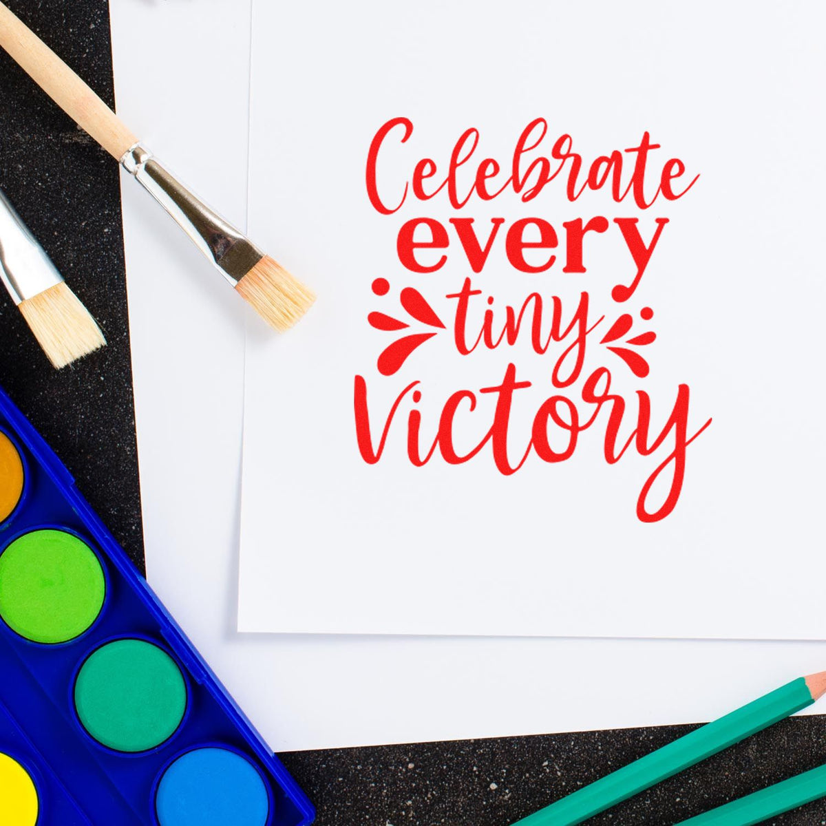Red "Celebrate every tiny victory" stamp/seal design with watercolor brushes, blue paint palette, and pencils on white paper.