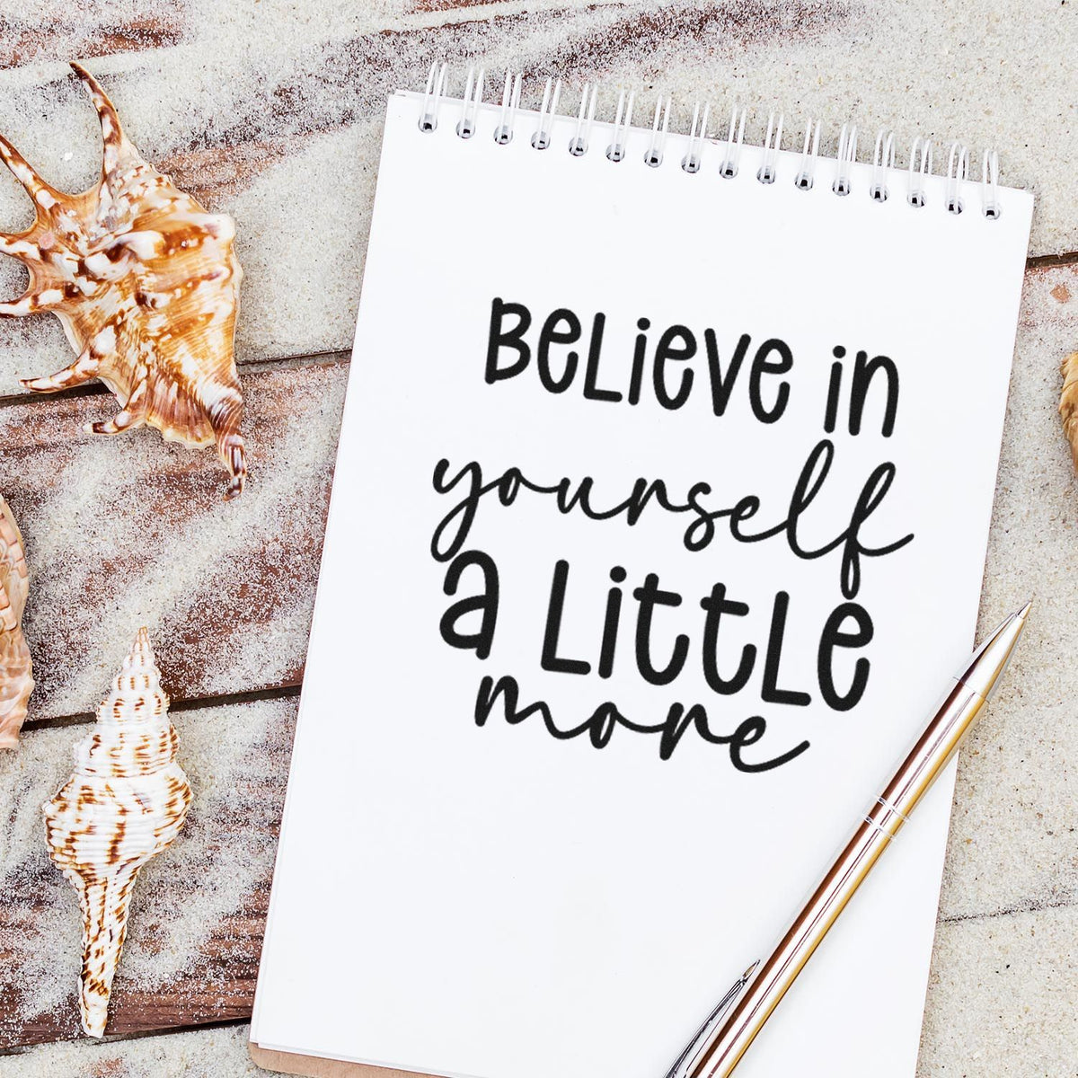 Custom stamp and seal on white spiral notebook reading Believe in yourself a little more, with seashells and a gold pen.