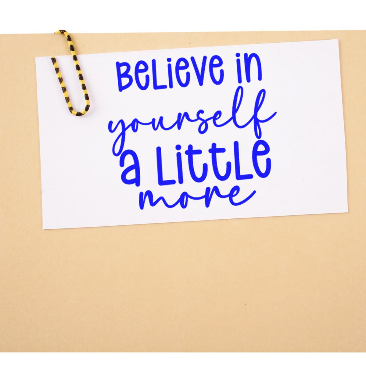 Blue stamp-style quote Believe in yourself a little more on white card clipped with yellow-black paper clip against beige background.