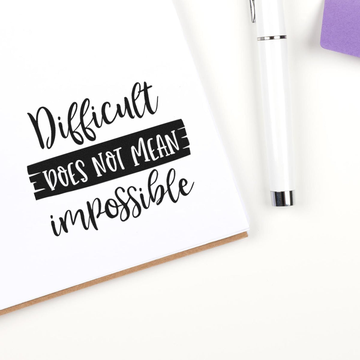 Black motivational stamp and white seal pen on white paper reading Difficult does not mean impossible, with a purple accent.