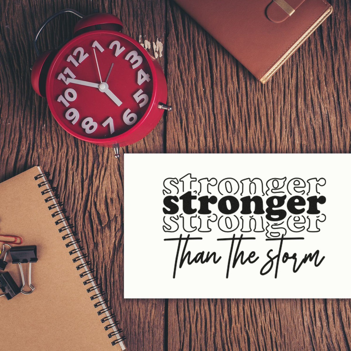 White stamp and seal design reading stronger than the storm beside a red alarm clock, notebook, and planner on wood.