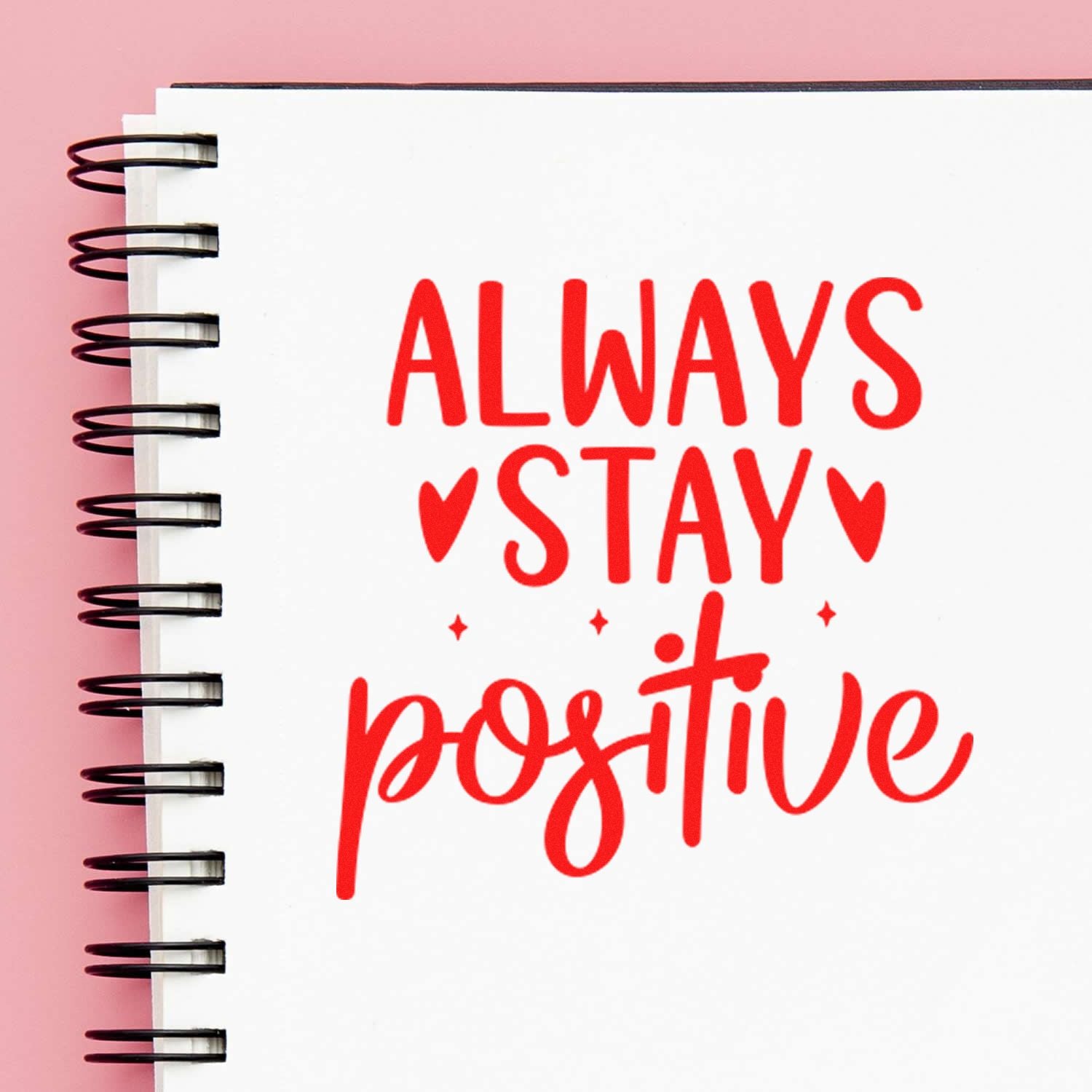 Red Always stay positive stamp and seal design on white spiral notebook with black rings, pink background, hearts and sparkles.