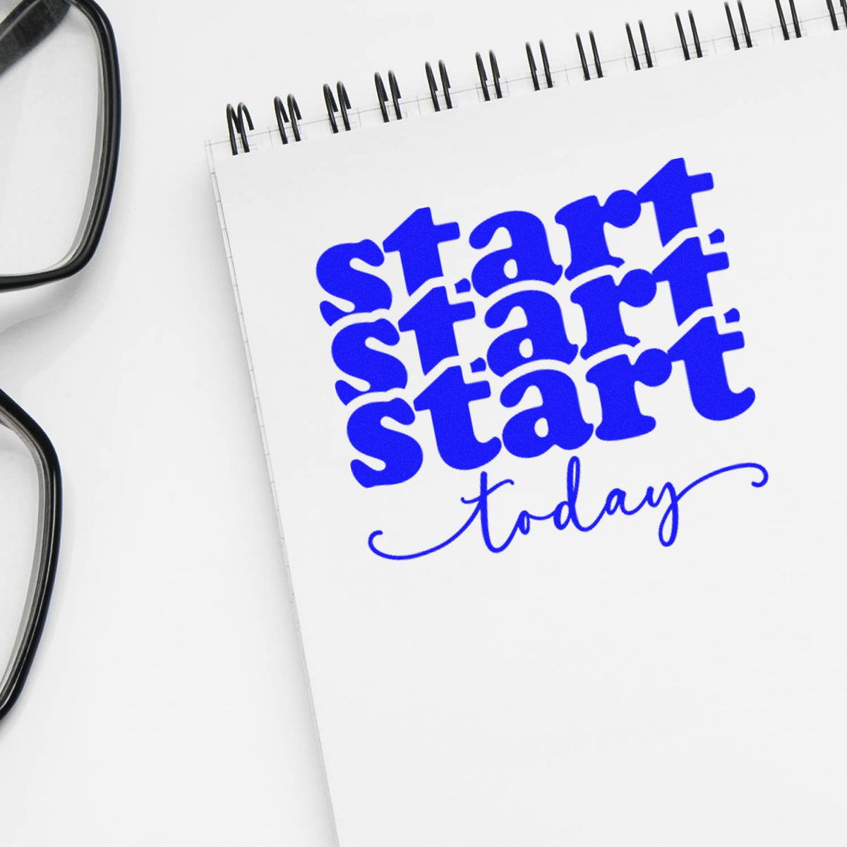 Blue custom stamp and seal reading start today on white spiral notebook beside eyeglasses.