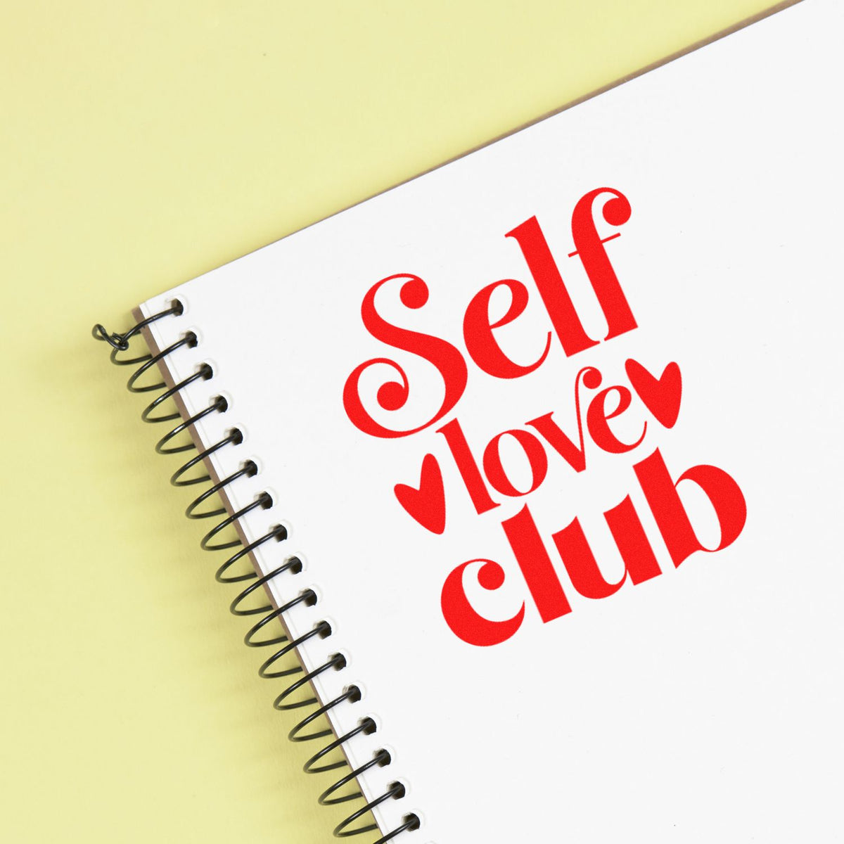 Custom stamp and seal design with red Self Love Club text and hearts on a white spiral notebook over pale yellow.