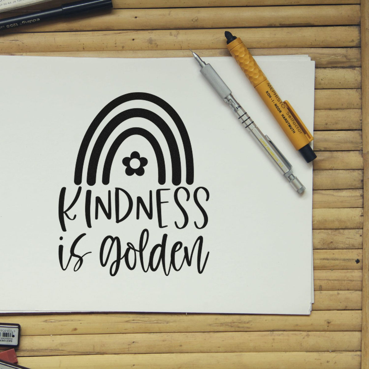 Black Kindness is Golden stamp on white paper with rainbow and flower, beside gold and silver seal tools on wood.
