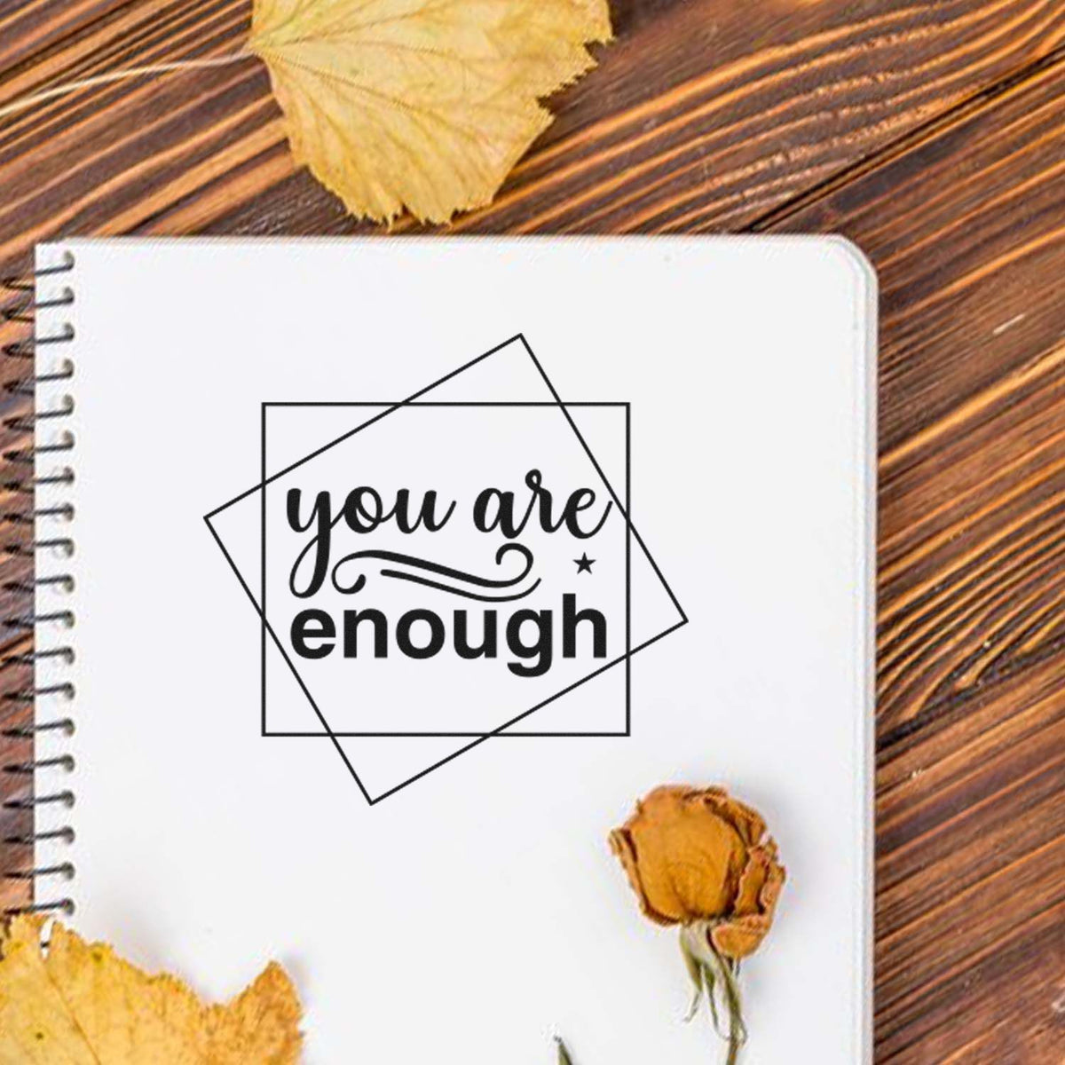 Black you are enough stamp seal on a white spiral notebook, with autumn leaves and a dried flower on a brown wood surface.