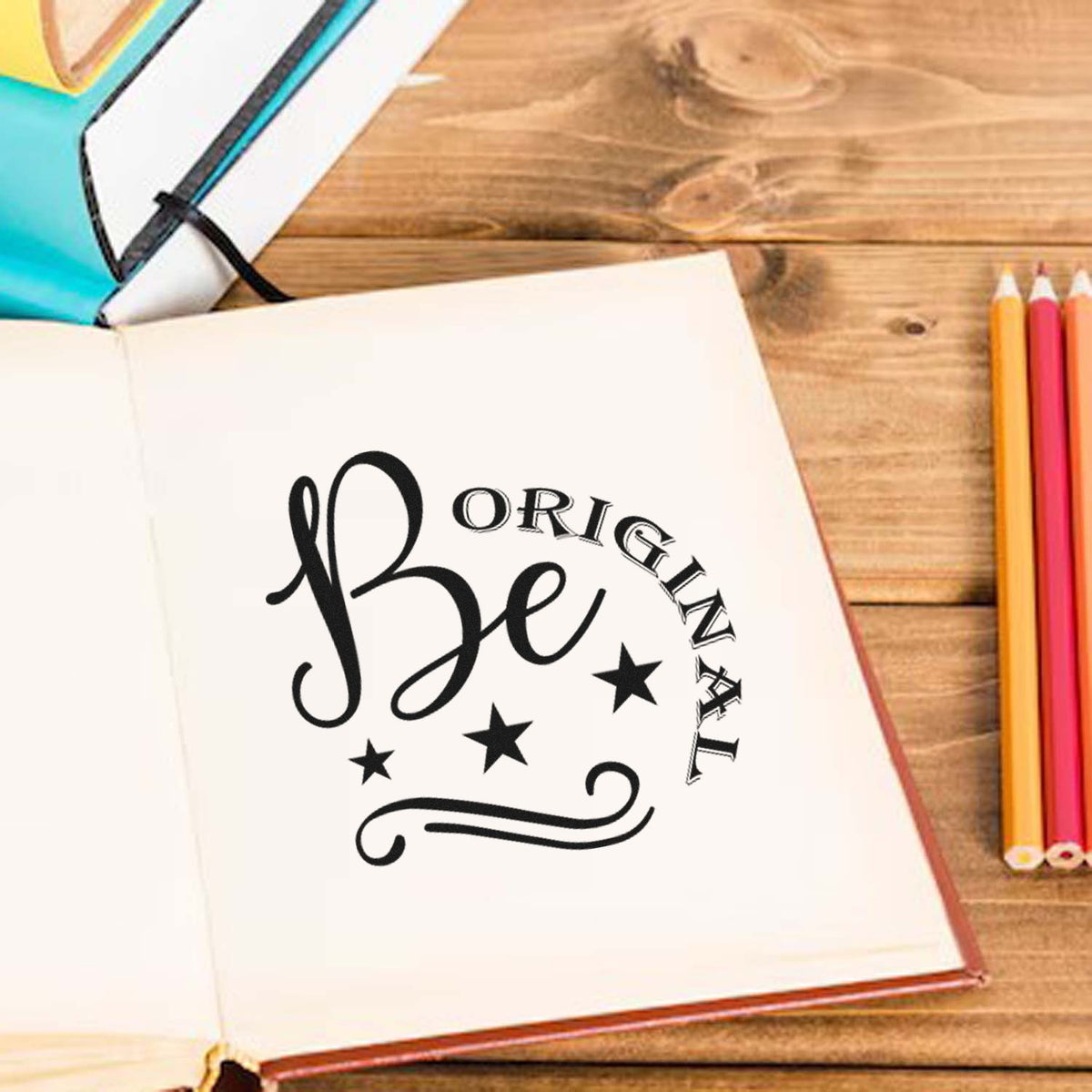 Black Be Original stamp seal design on cream notebook, with stars, beside colored pencils on rustic wood.