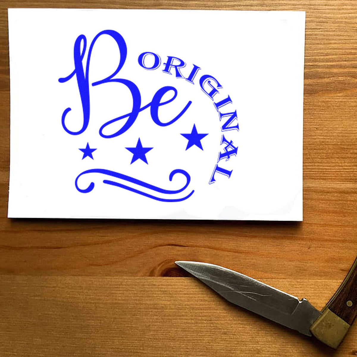 Blue Be Original rubber stamp seal with stars and swirls on a white card against a wood tabletop.