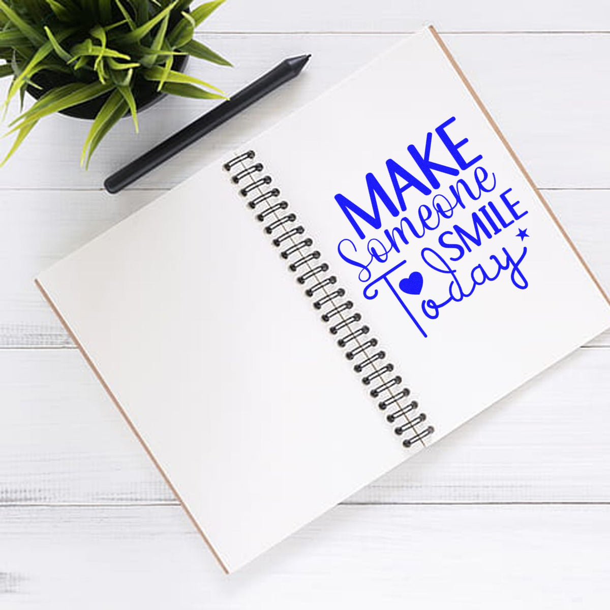 Blue custom stamp and seal design on spiral notebook reading MAKE someone SMILE Today with pen and plant.
