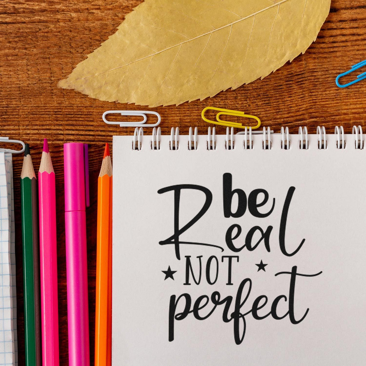 Decorative stamp/seal reading Be Real Not Perfect on a notepad, with colorful pencils, paper clips, and a dried leaf.