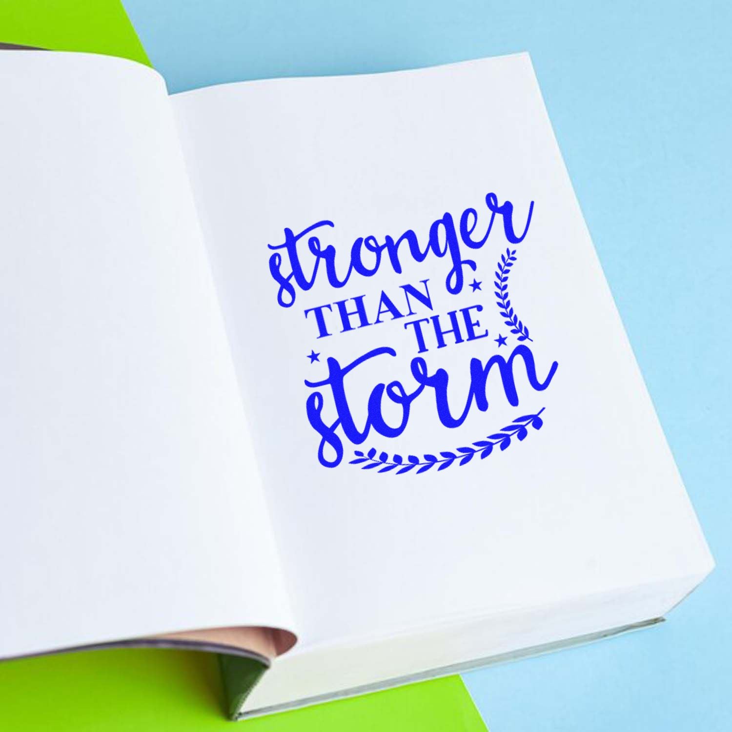 Blue Stronger Than the Storm stamp and seal on an open white notebook, with laurel accents on a light blue and lime background.