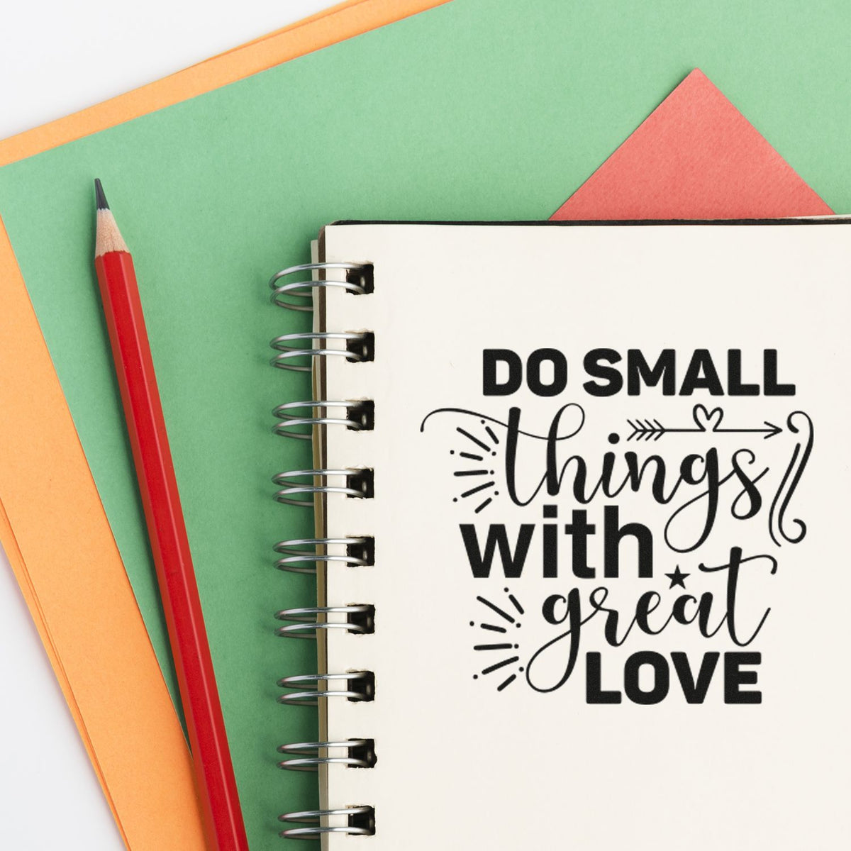 Custom stamp and seal design on a spiral notebook with Do small things with great love in black, plus a red pencil and green-orange paper.