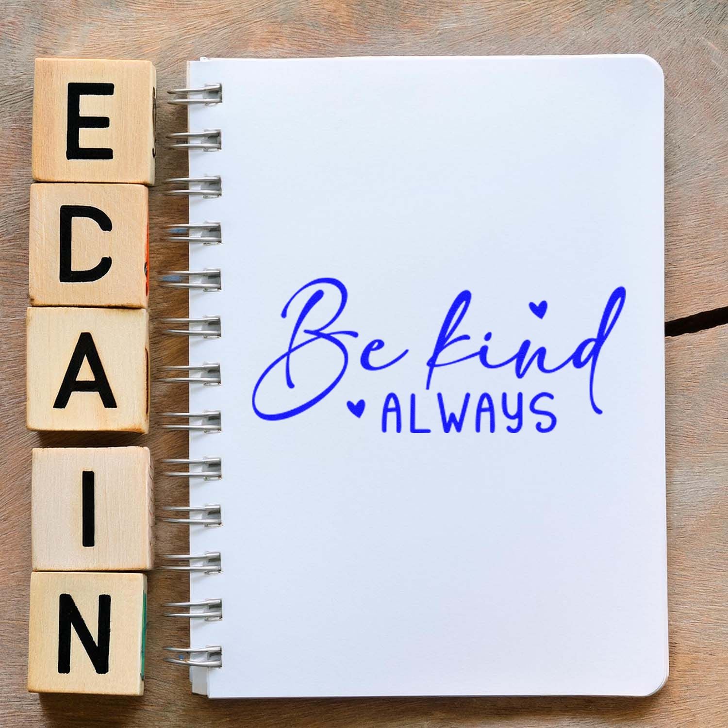 Wooden alphabet stamp blocks beside a spiral notebook stamped in blue with Be kind Always and hearts.