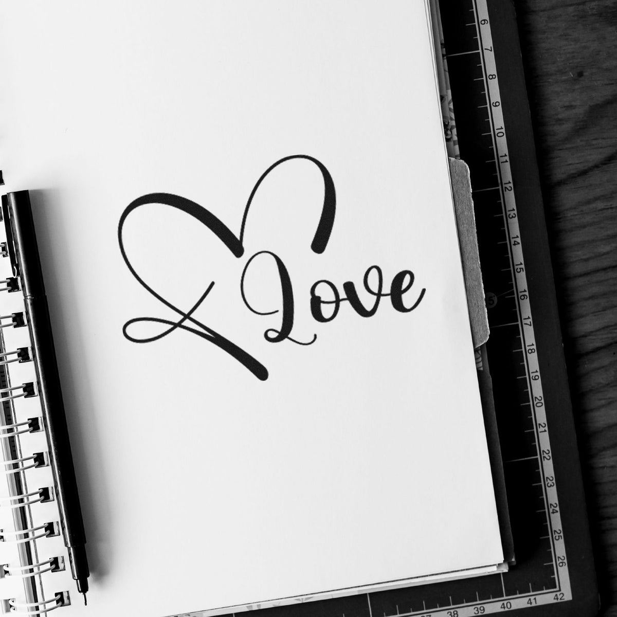 Black love seal stamp with heart outline on a white notebook page, beside a pen and ruler.