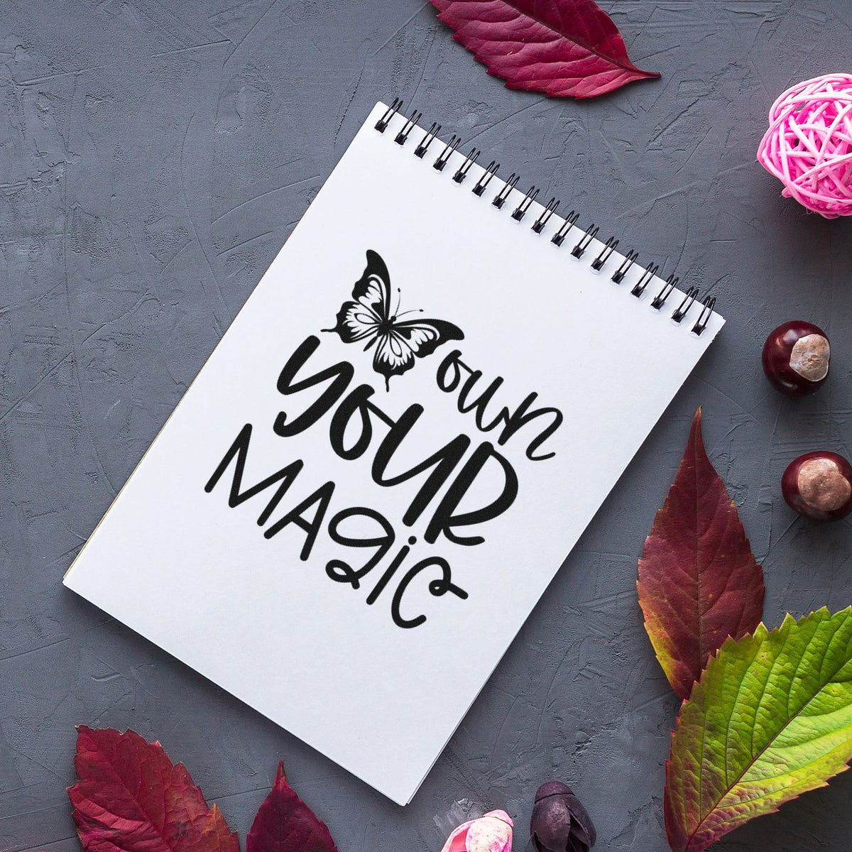 Black butterfly stamp and seal reading Your Magic on a white spiral notebook, with burgundy leaves and pink accents.