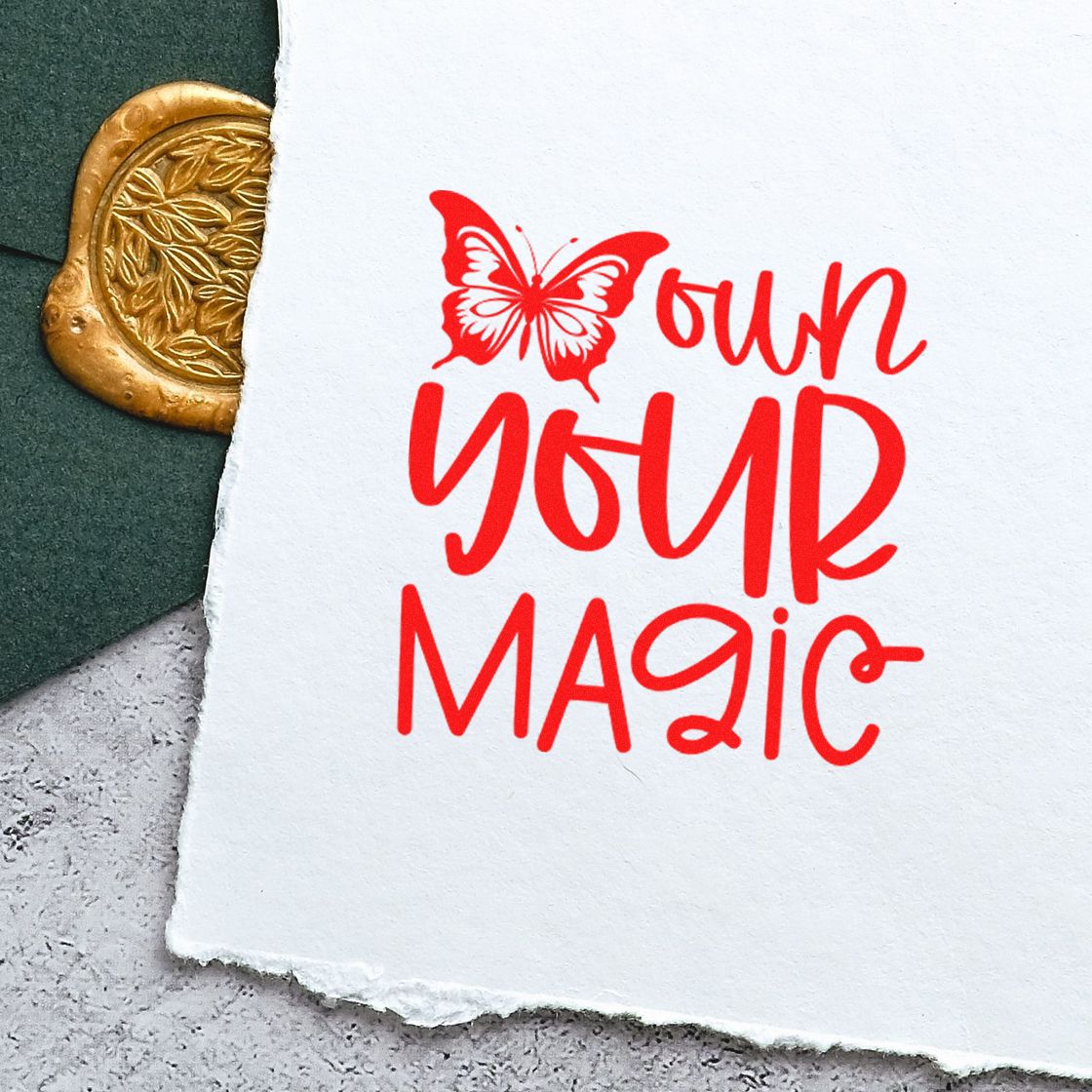 Red butterfly stamp reading own your magic beside a gold embossed wax seal on white paper.