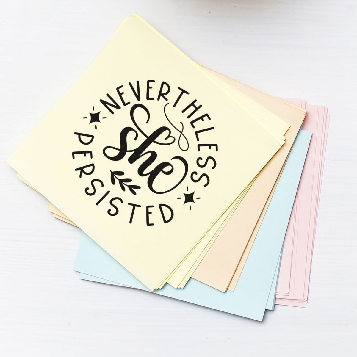 Stack of pastel stamp and seal cards with black She Persisted design on cream, blue, pink, and beige sheets.