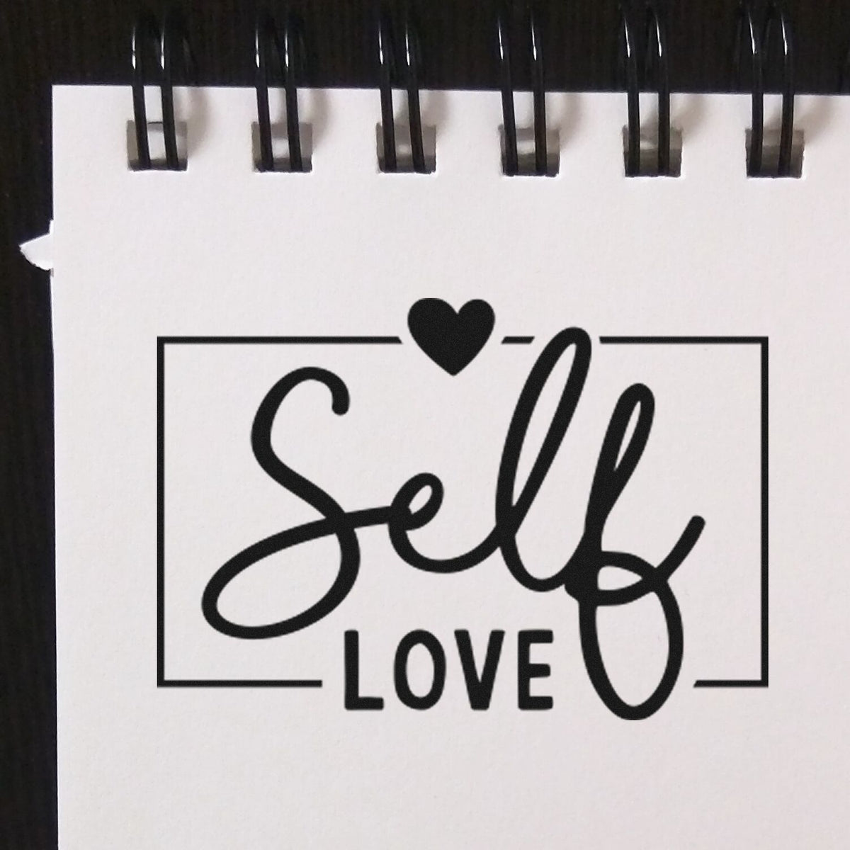 Black self-love stamp and seal design with heart on white spiral-bound paper.