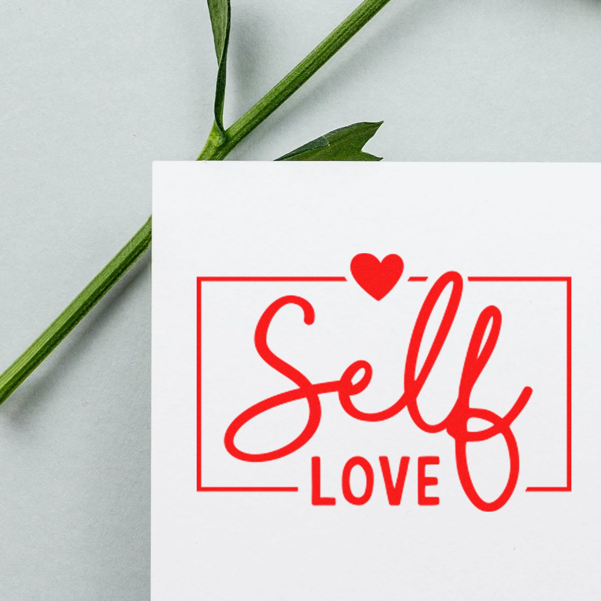 Red Self Love stamp with heart on a white card, beside a green stem; decorative stamp and seal product.