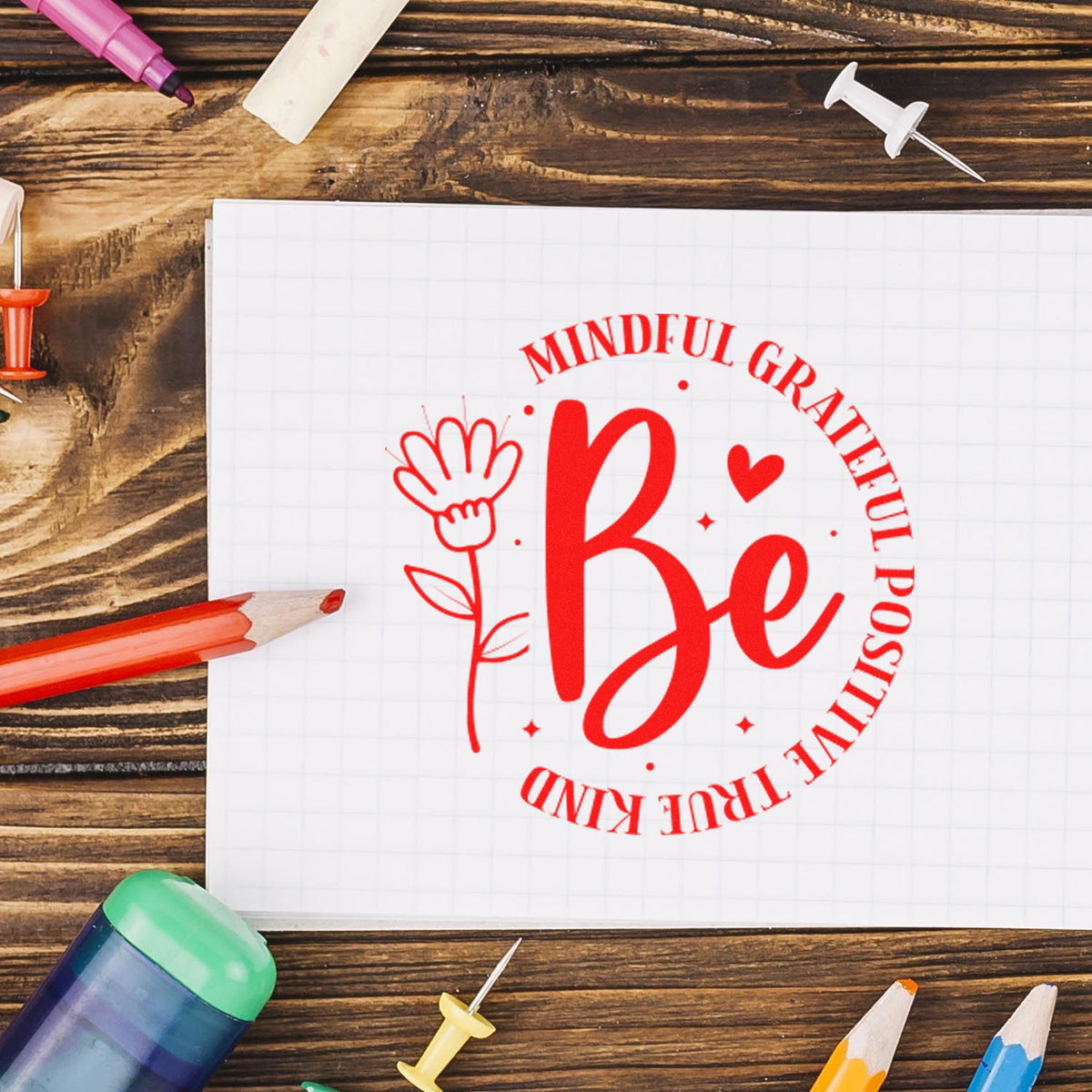 Red mindfulness Be stamp seal on white grid paper, surrounded by colorful pencils and push pins on a rustic wood table