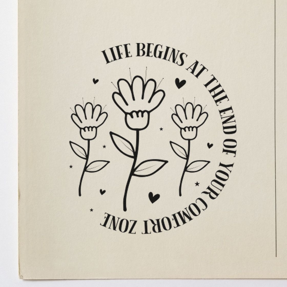 Black circular rubber stamp seal with three flowers and hearts on cream paper, reading Life begins at the end of your comfort zone.