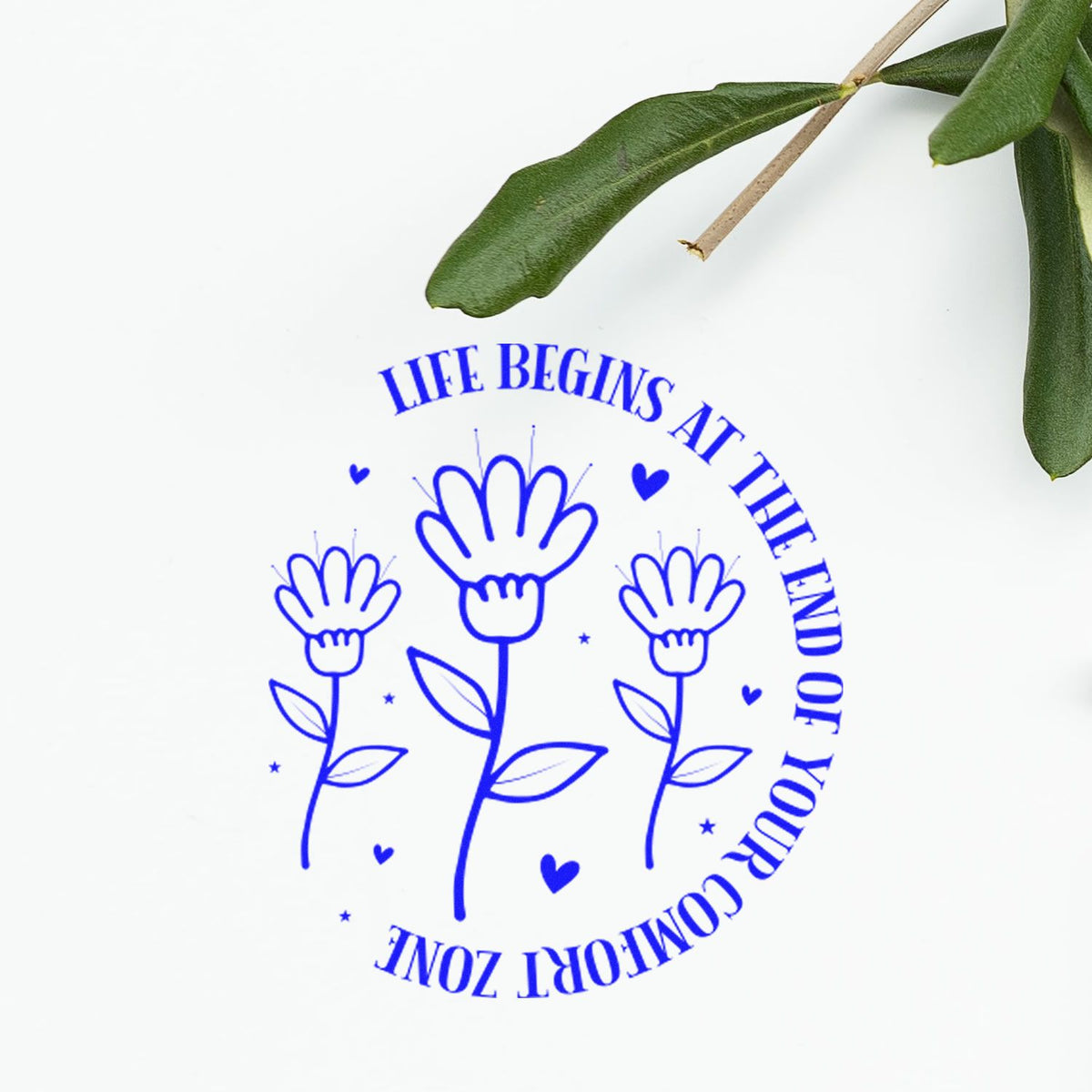 Blue floral rubber stamp seal with Life begins at the end of your comfort zone text on white, accented by green leaves.