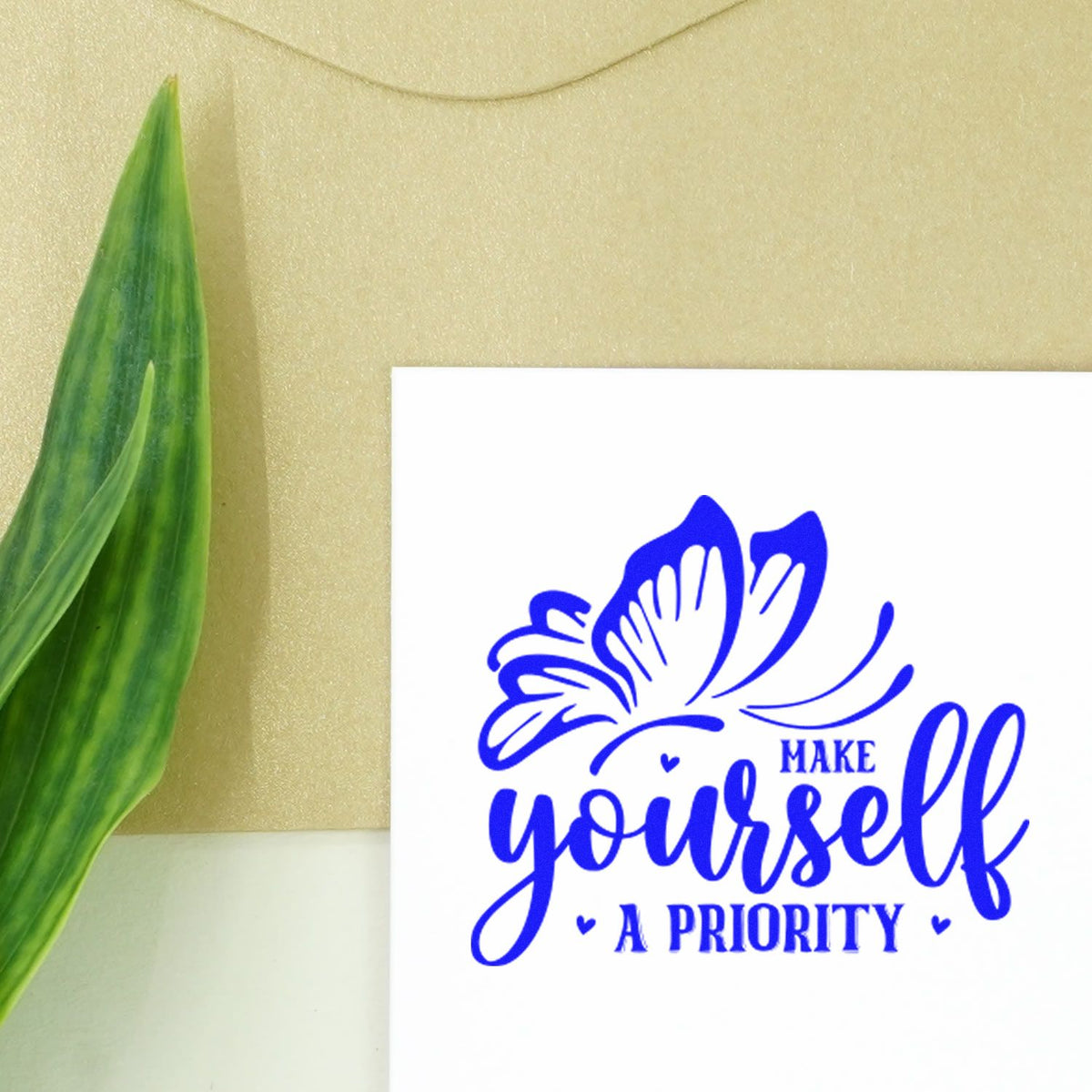 Blue butterfly custom stamp and seal reading Make yourself a priority on white card, beside a green leaf.