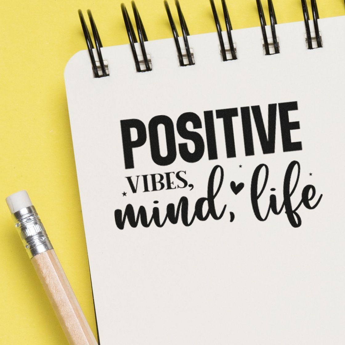 Decorative stamp/seal reading POSITIVE vibes, mind, life on a white spiral notebook with a yellow background and pencil.