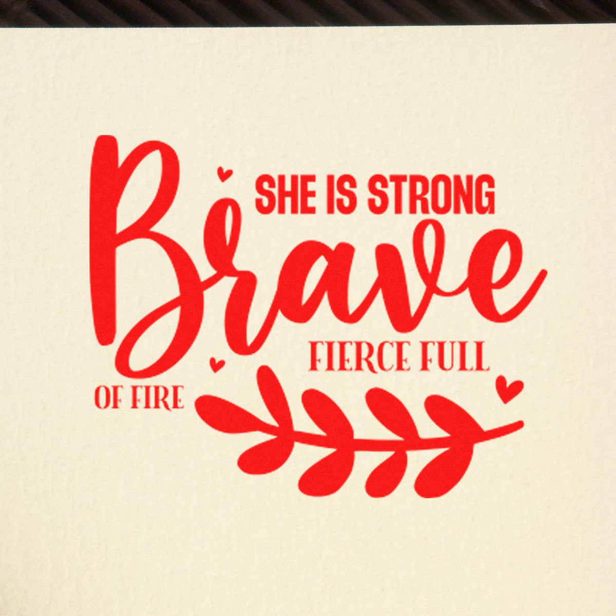 Red motivational stamp design with She is strong, brave, fierce, full of fire in bright red on cream, plus hearts and leafy branch.