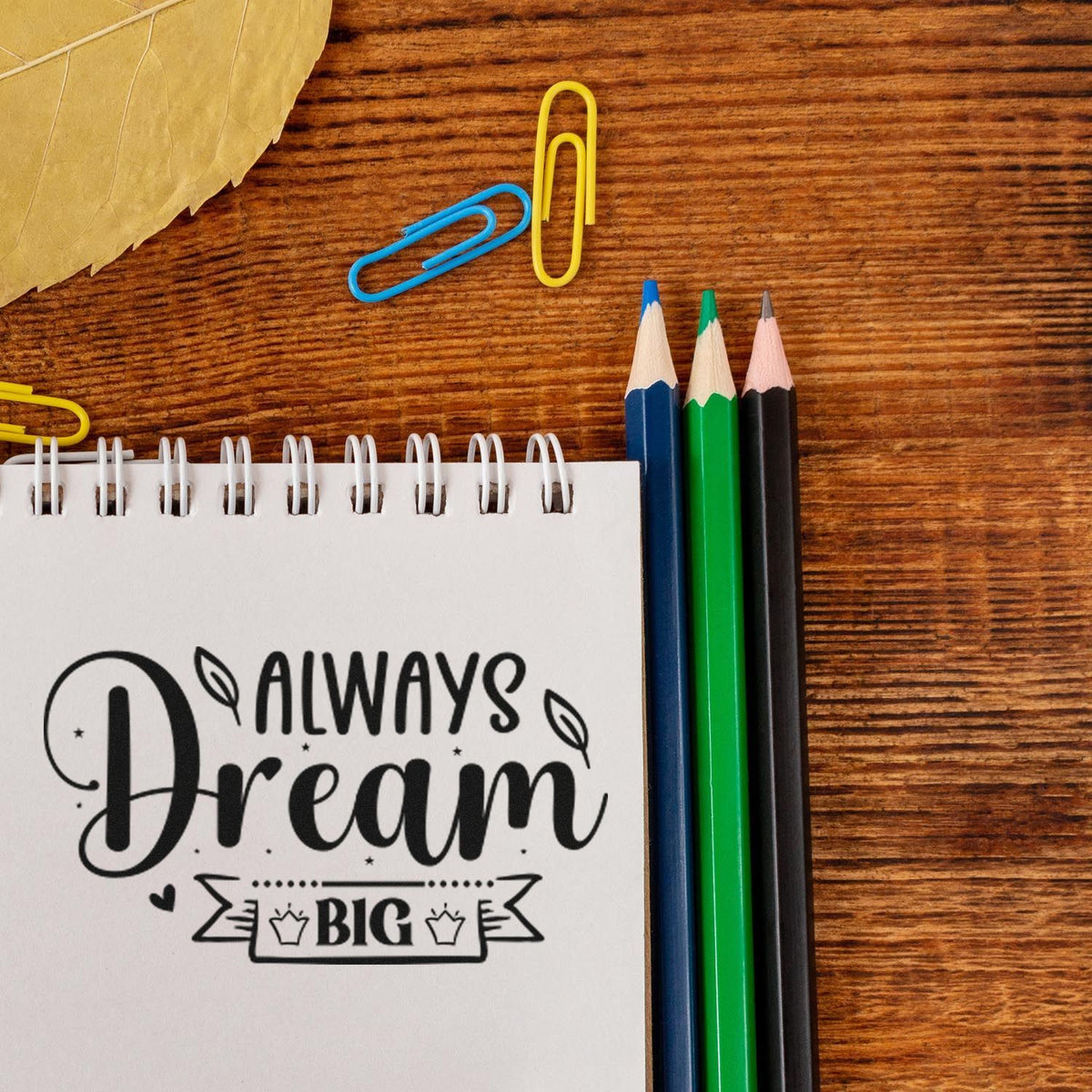 Black Always Dream Big stamp and seal on a white notepad, with blue, green and black pencils and yellow-blue paper clips.