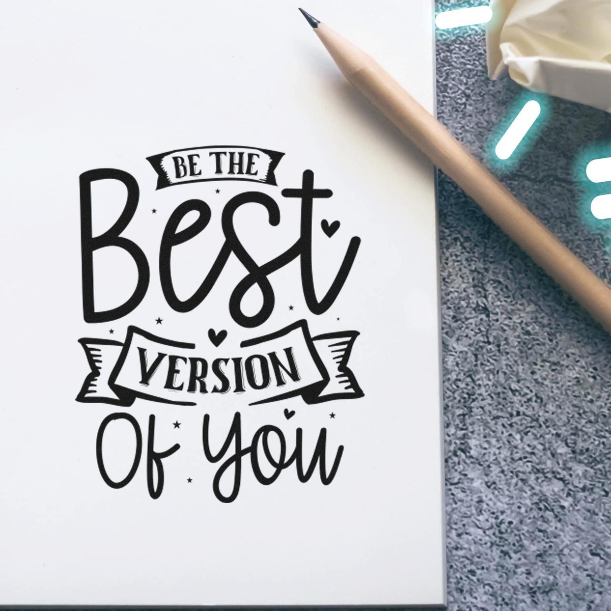 Black decorative stamp and seal reading Be the Best Version of You on white paper beside a wooden pencil.