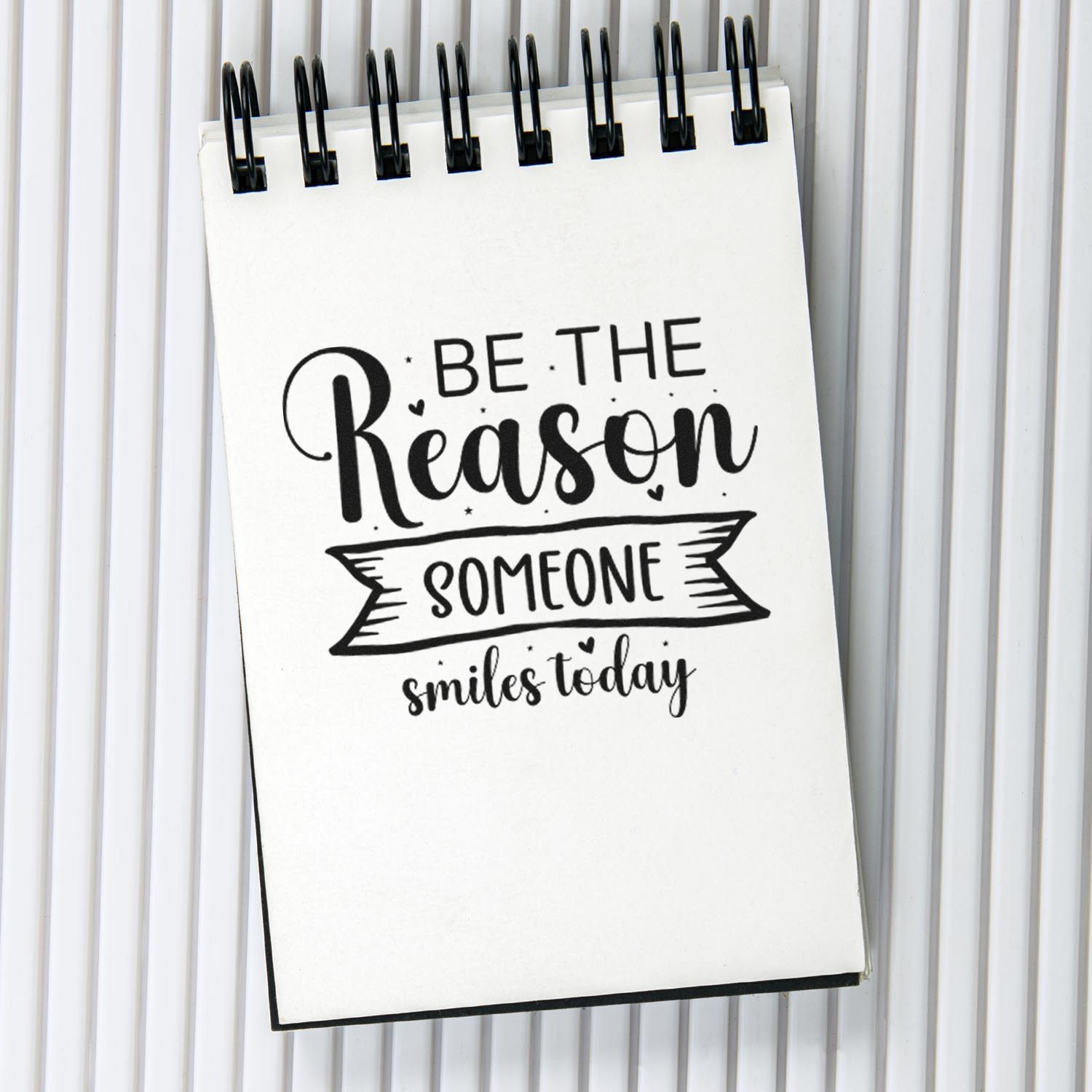 Black custom stamp seal on white notepad reading Be the reason someone smiles today.