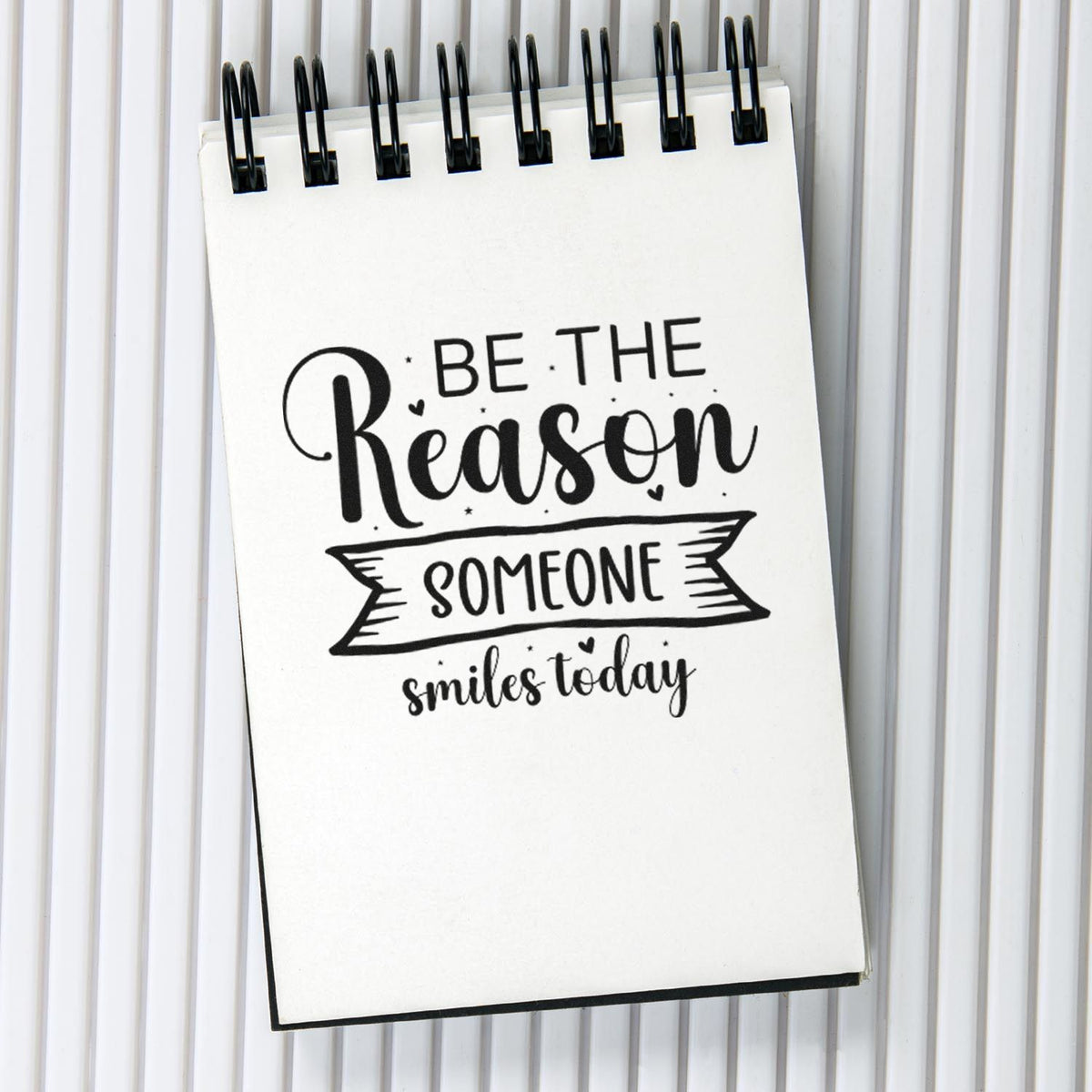 Black rubber stamp on white spiral notepad reading Be the reason someone smiles today in bold script.