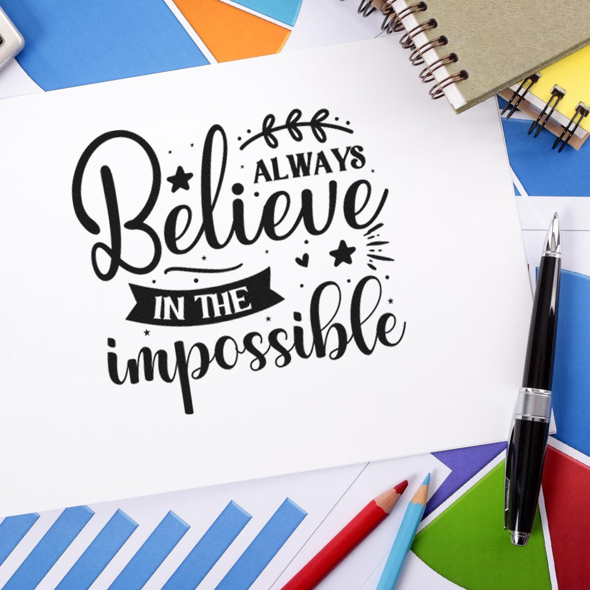 Black stamp and seal reading Always Believe in the Impossible on white paper, with colorful envelopes and pens.