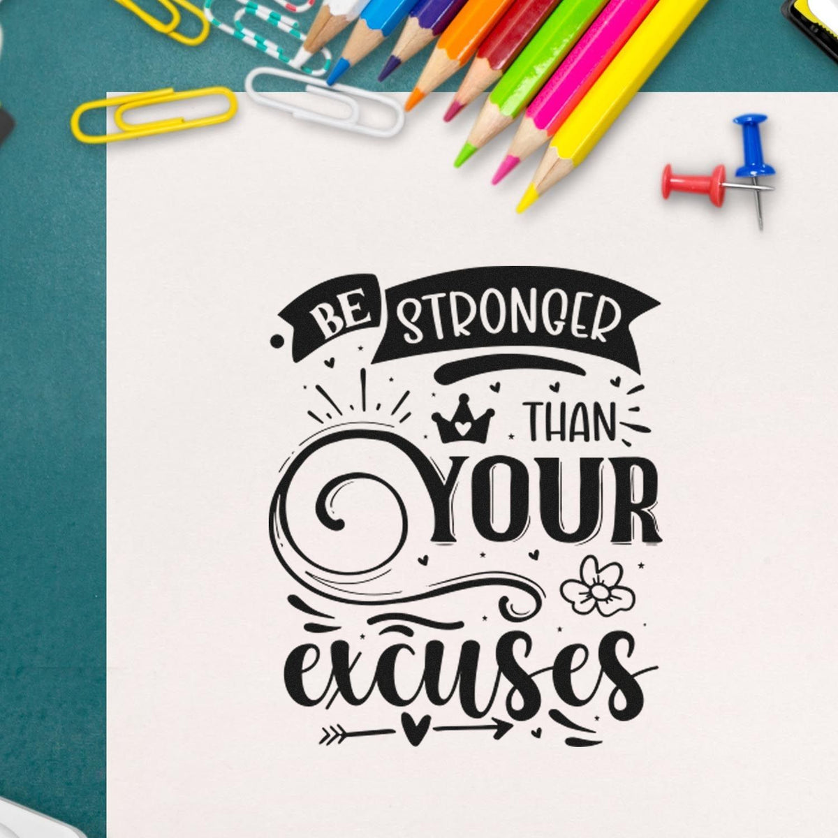 Black motivational stamp/seal reading Be Stronger Than Your Excuses on white paper, with colorful pencils and push pins.