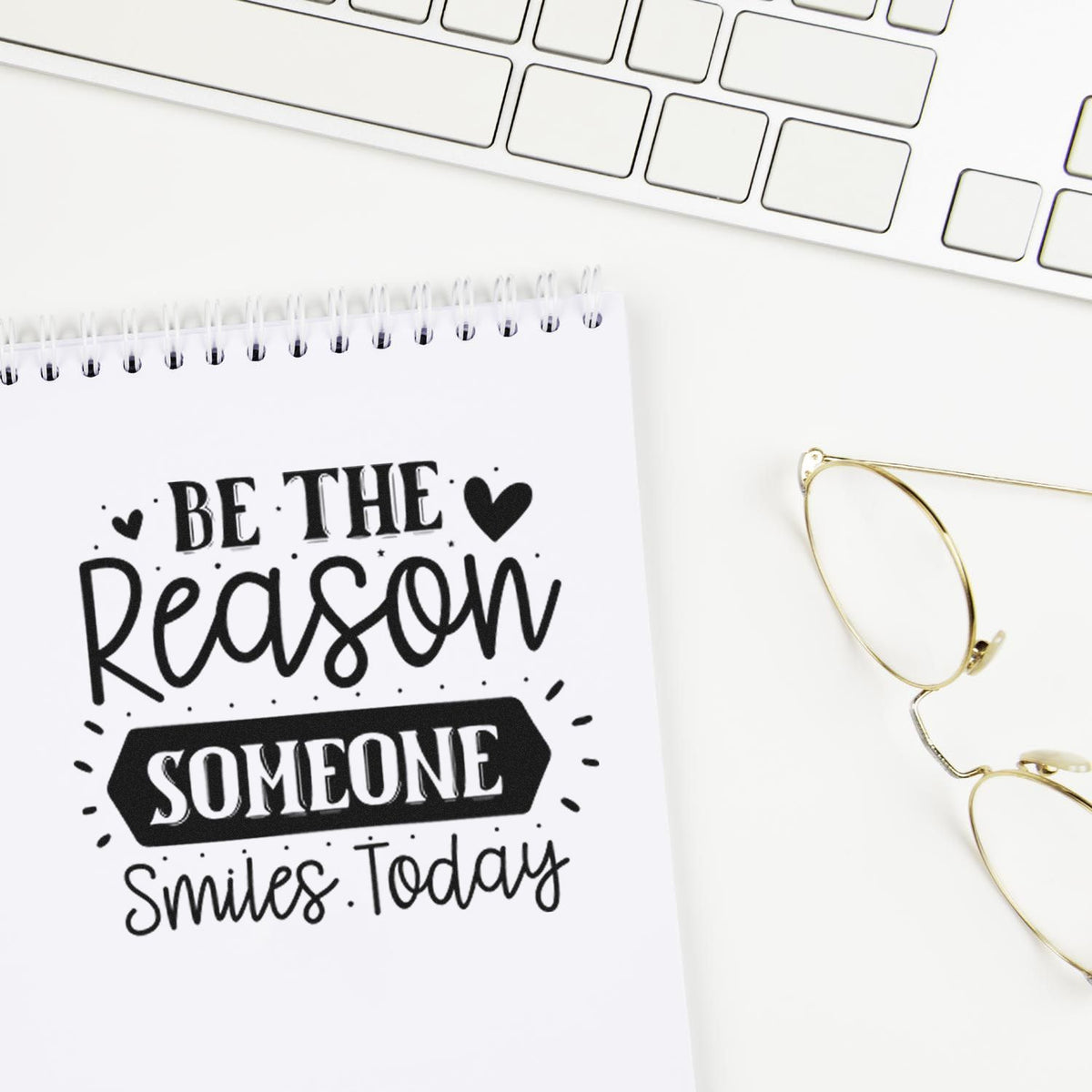 Black stamp and seal design on white notepad reading Be the reason someone smiles today, beside gold glasses and keyboard.