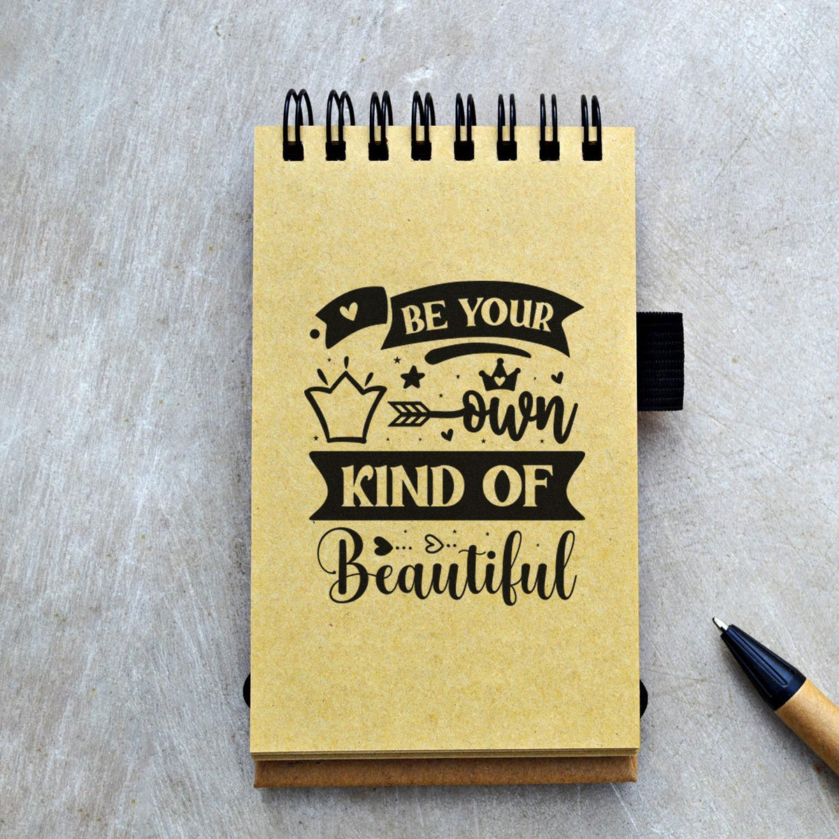 Yellow spiral notebook featuring a black custom stamp seal design reading Be Your Own Kind of Beautiful with crown and heart accents