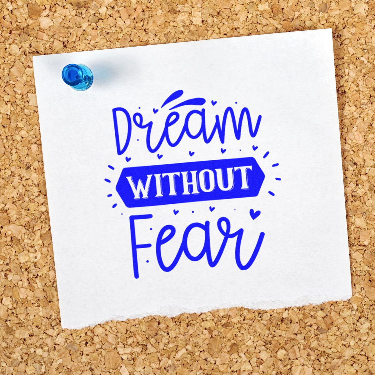 Blue rubber stamp seal reading Dream Without Fear on white paper, pinned with a blue tack against tan corkboard.