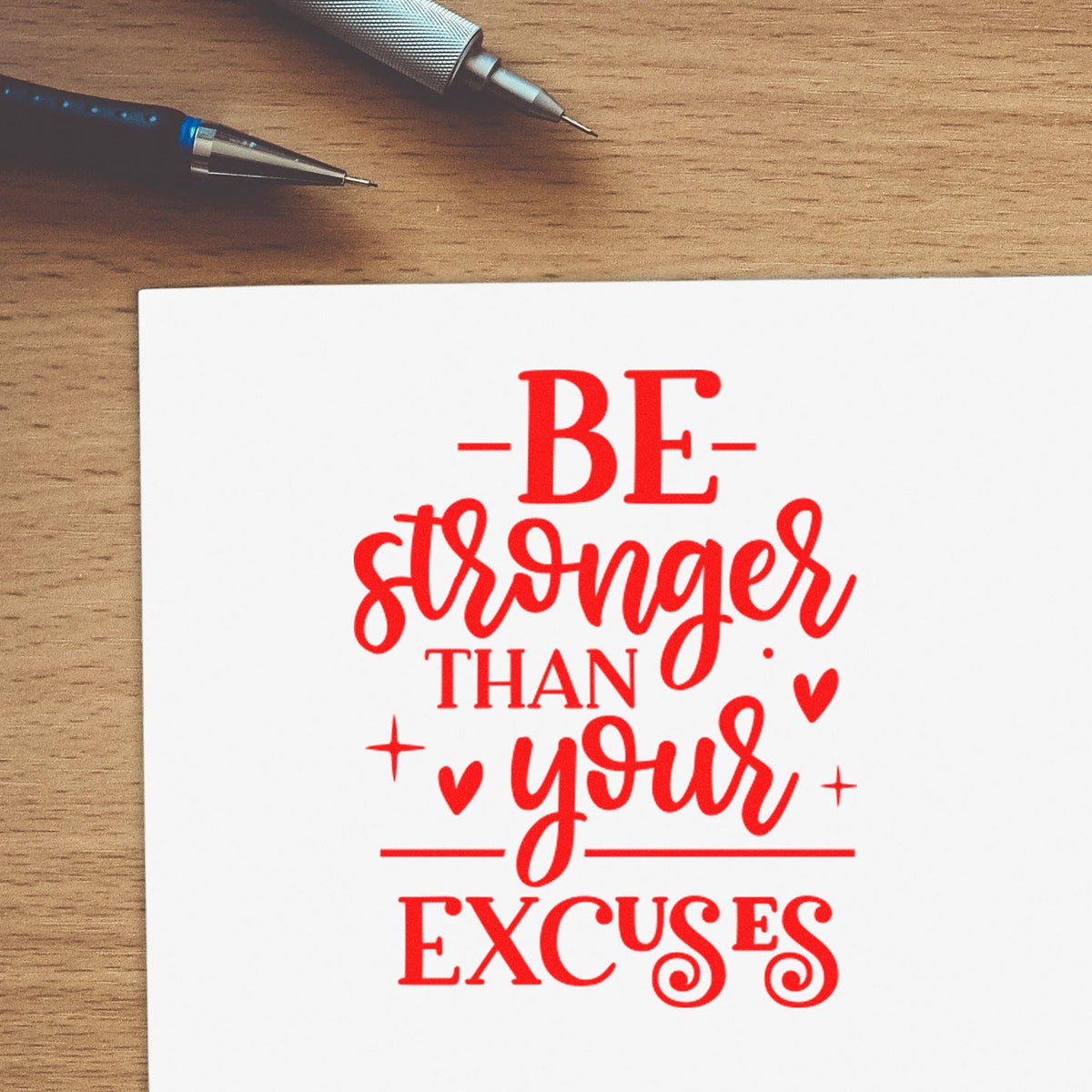 Red custom stamp/seal reading Be stronger than your excuses on white card, with two pens on a wood desk.