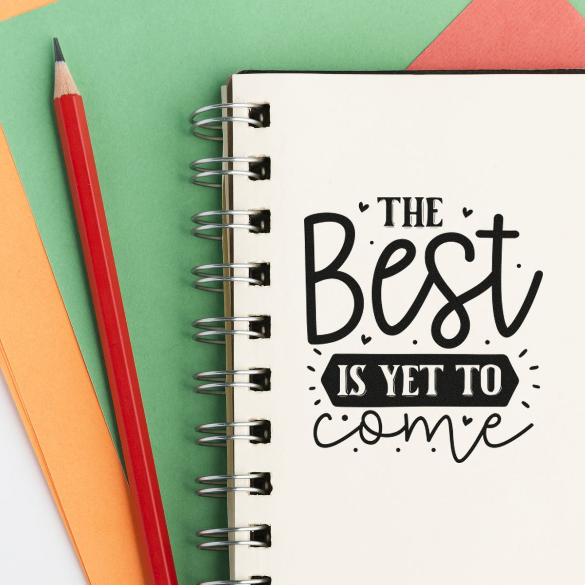 Black motivational stamp design on a spiral notebook, with a red pencil and green, orange, and coral paper background.