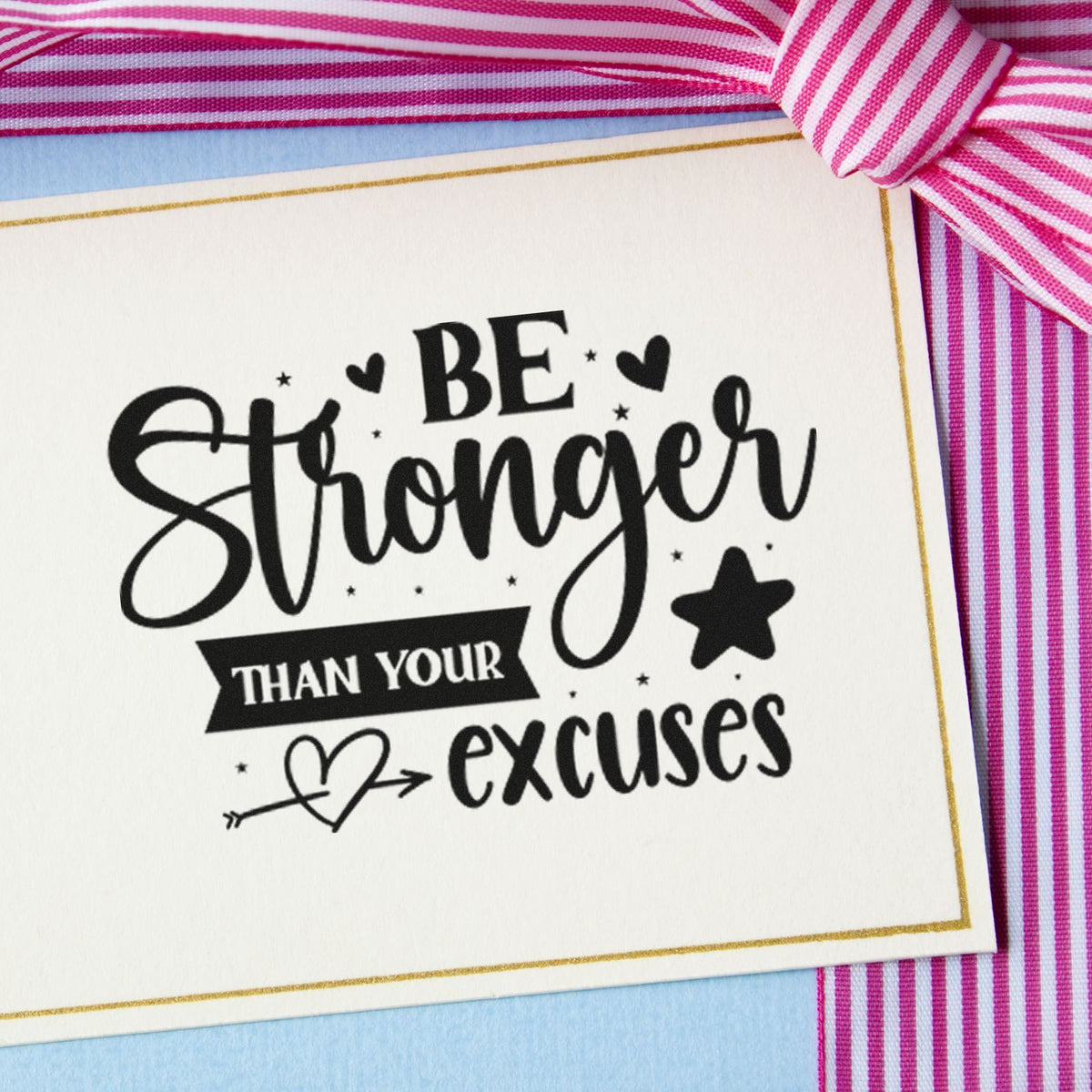 Black motivational stamp and seal reading Be Stronger than your excuses, with hearts, stars, gold border, and pink striped ribbon on blue background.