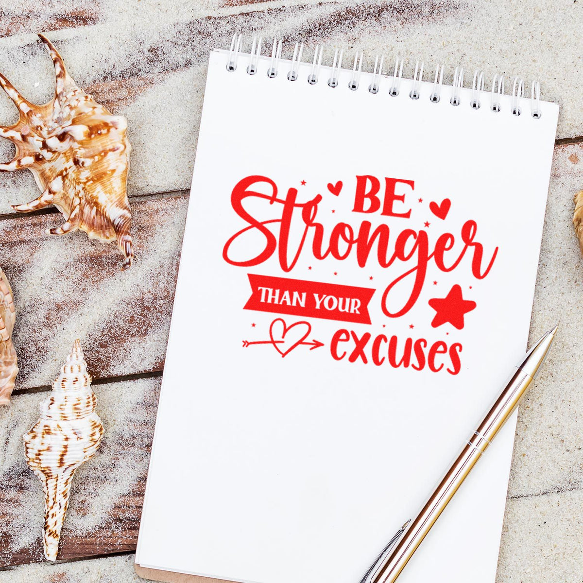 Red motivational stamp seal on a white spiral notebook, with seashells and a gold pen on a sandy wood background.