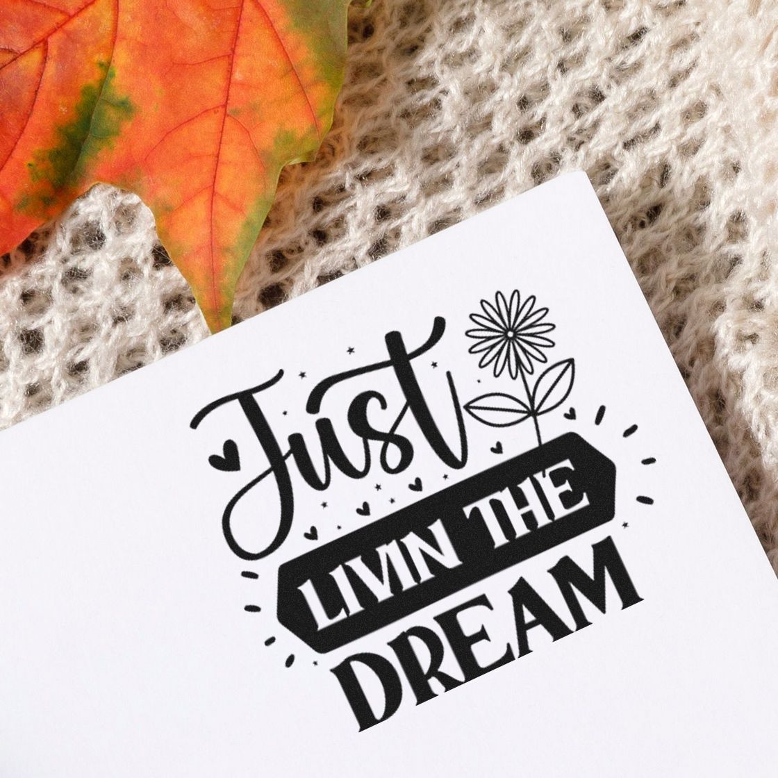 Black custom stamp seal on white paper reading Just Livin the Dream, with hearts, flower, and orange leaf on knit background.