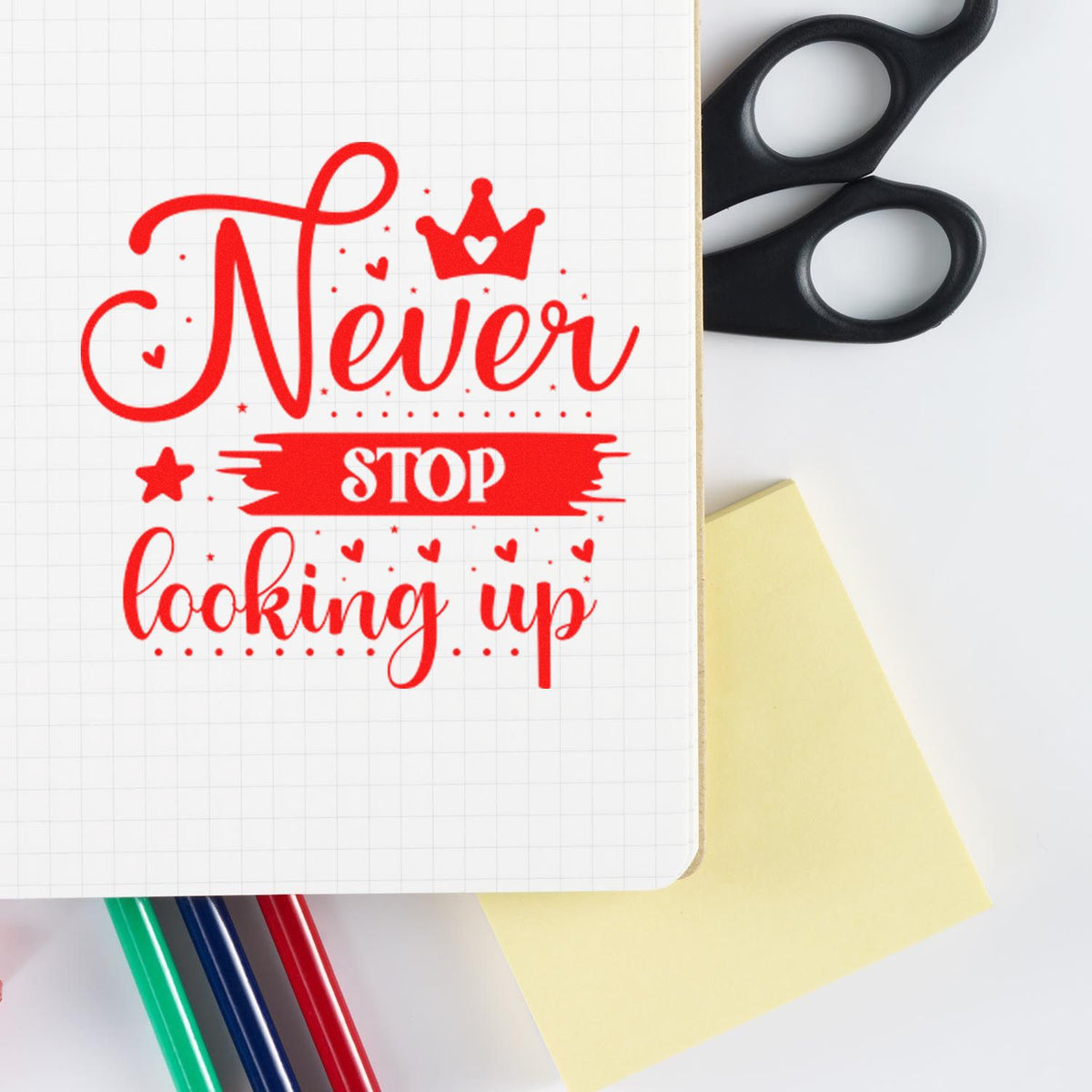 Red Never Stop Looking Up stamp and seal design on graph paper, with black scissors, yellow note, and colored pens.