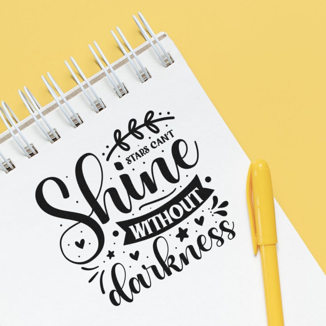 Black stamp and seal design reading Shine without darkness on a white spiral notebook with a yellow pen on a yellow background.